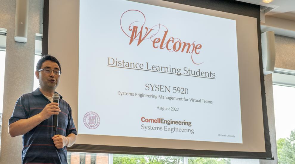 Oliver Gao stands in front of a screen that reads "Welcome Back Distance Learning Students"