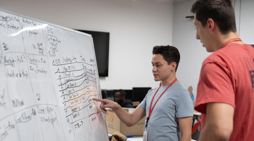 Two students discuss a plan that they have listed on a whiteboard in the lab.